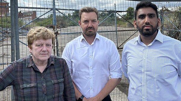 Councillors Sandra Gidley, Geoff Cooper and Nik Daas stand infront of a metal fence, behind the fence is a concrete surface overgrown with shrubbery 